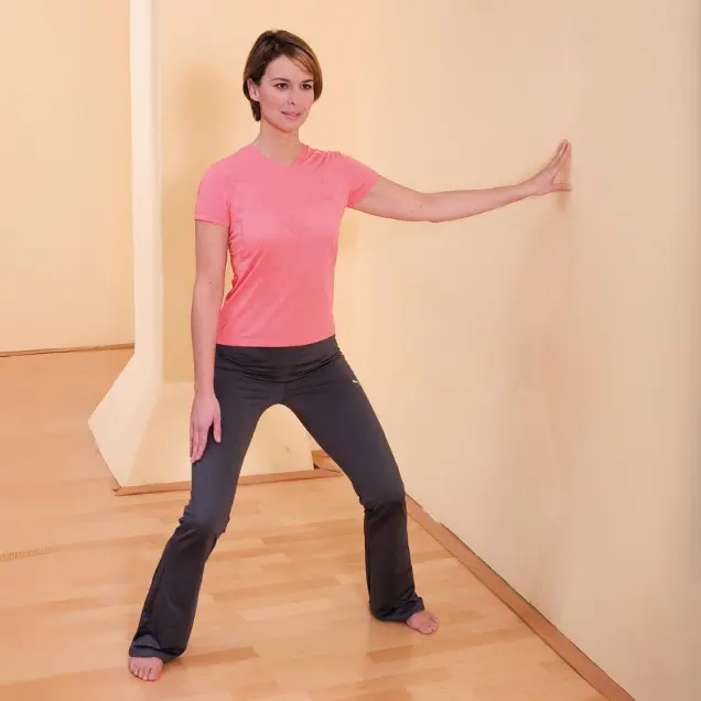 Frau steht mit leicht gebeugten Knien neben einer Wand und stützt sich ab.